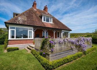 exterior of a chalet bungalow with a large wisteria-clad wooden deck, all surrounded by open countryside.