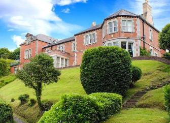 A large brick country house overlooks a sloping mature garden.