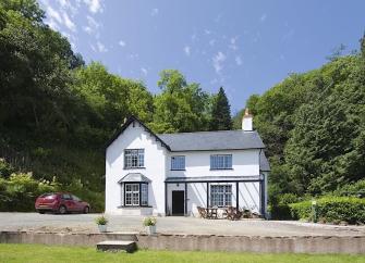 A 2-storey Exmoor holiday cottage stands in front of a bank of mature trees.