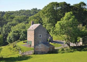 A tall stone barn conversion and annexe surrounded by a lawn, open fields and mature woods.