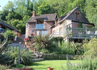A holiiday cottage with a large raised deck is surrounded by trees and mature gardens.