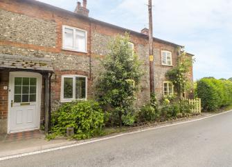 A stone-built Dorset holiday cottage behind a row of mature shrubs overlooks an empty country lane.