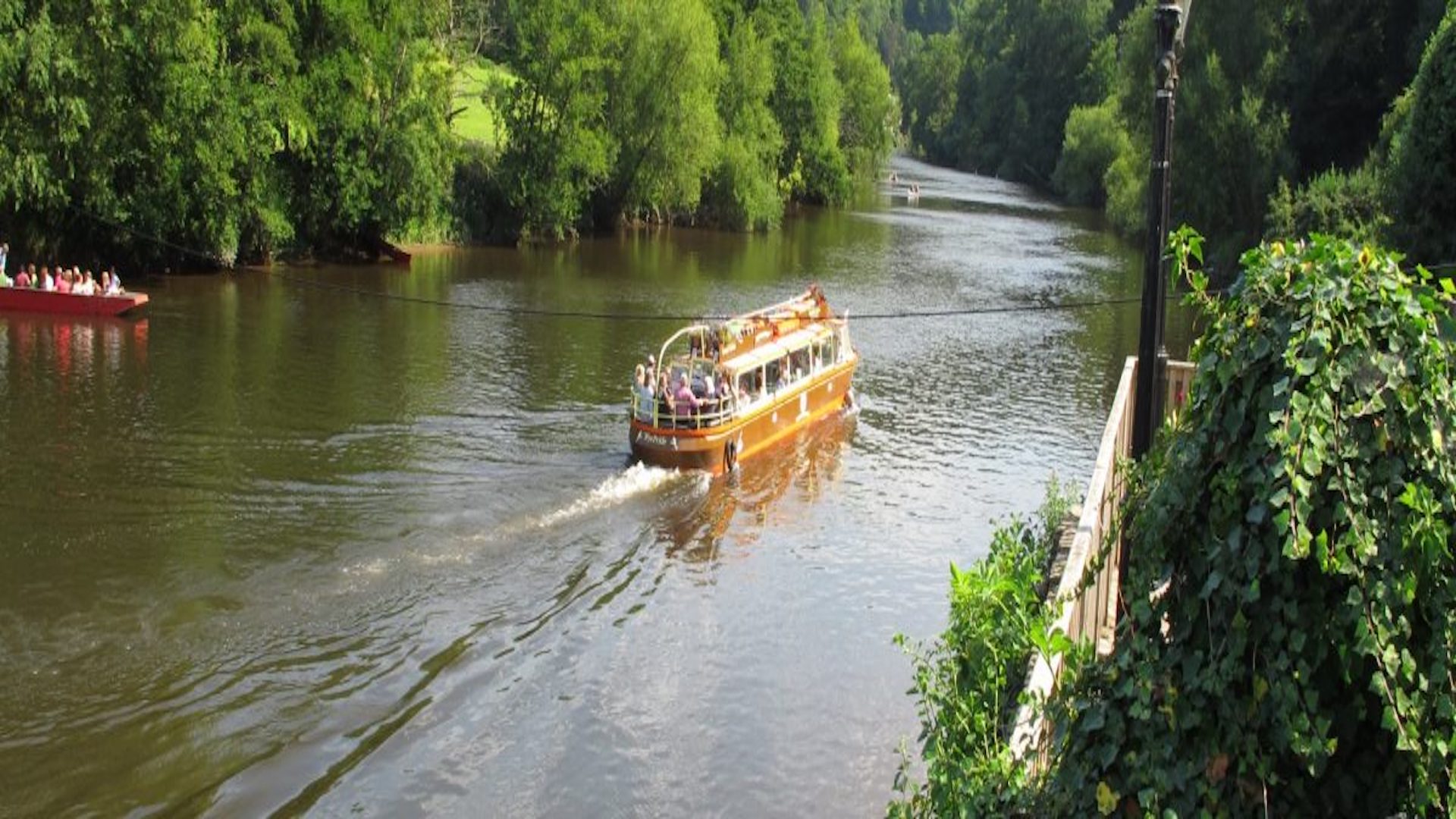 A pleasure boat sails slowly along a wide section of the River Wye in Herefordshire. Trees line both banks.