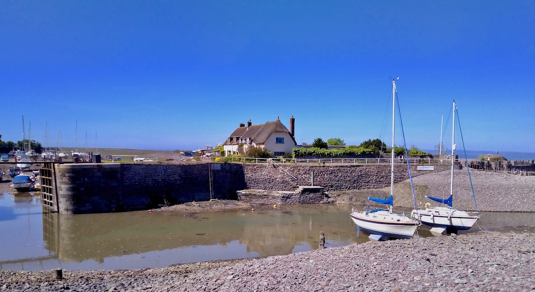 A small harbourside at low tide with two yachts moored on the pebble beache. Behind them is a large thatched holiday cottage.