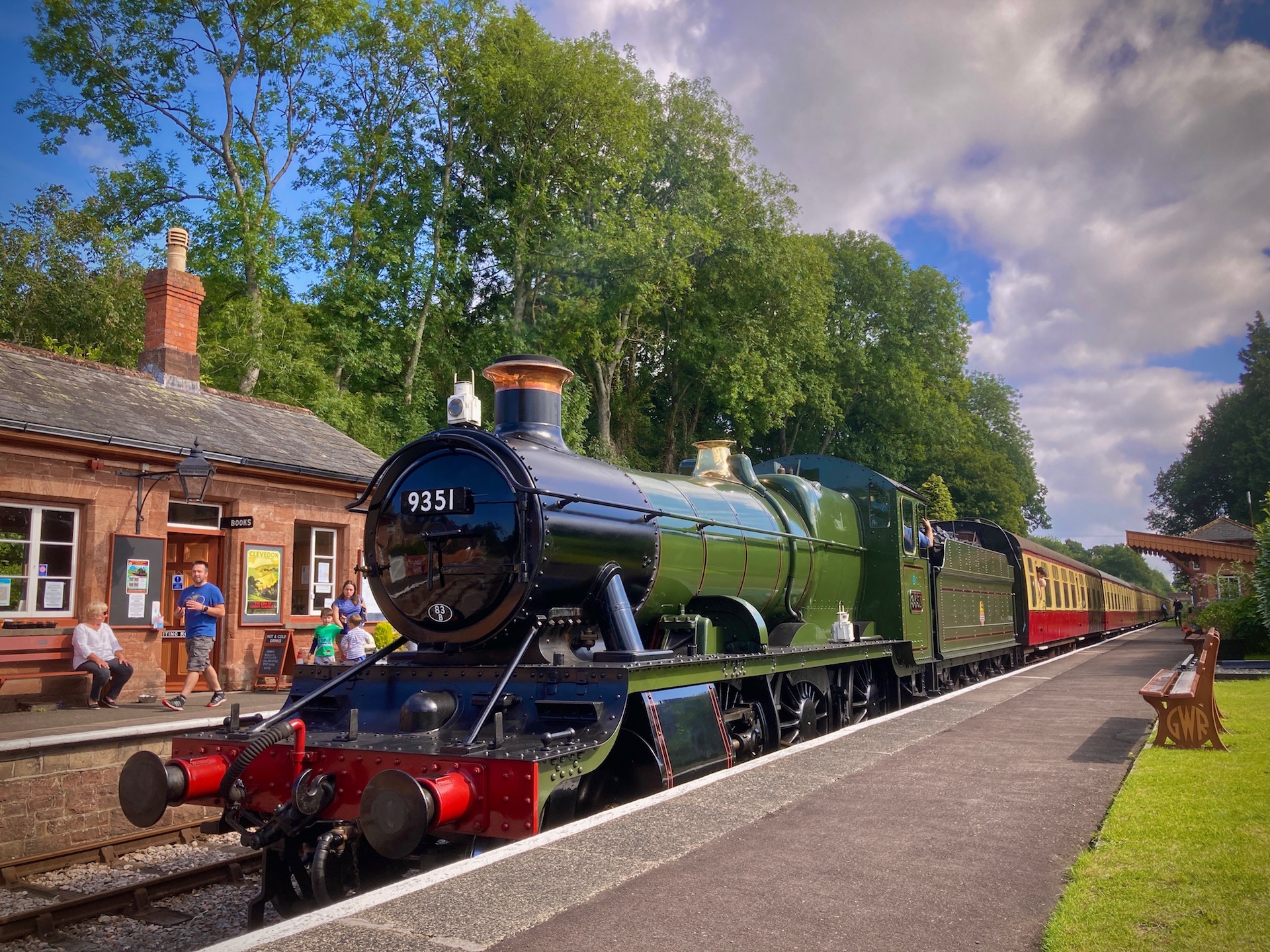 A large steam engines pulling a long line of carriages pauses at Crowcombe station in Somerset.