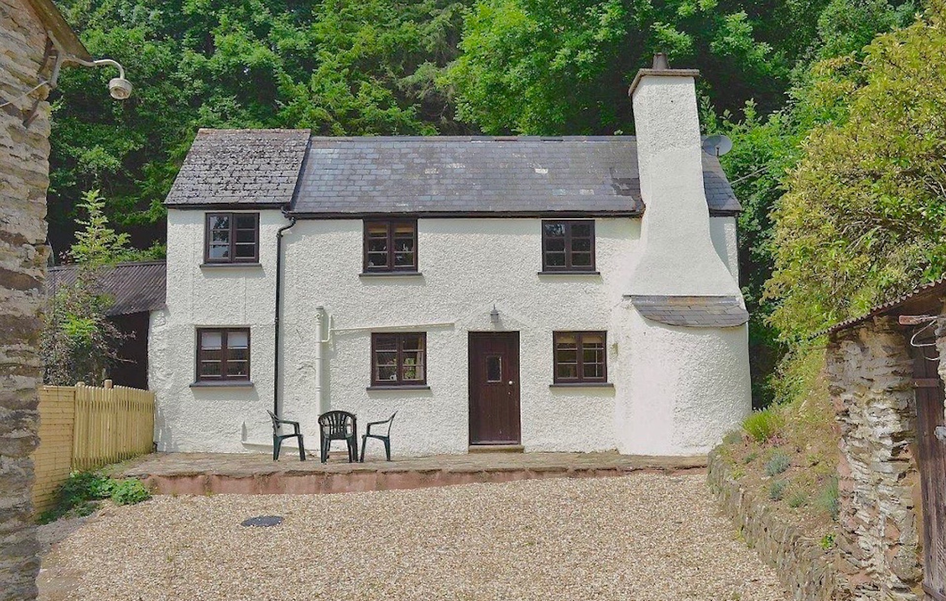 Exterior of a slate-roofed Exmoor holiday cottage with a large chimney and bread oven stack, overlooked by tall woodland trees..