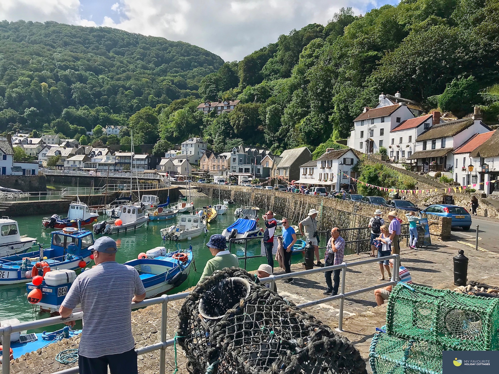 Lynmouth harbour full of pleasure and fishing boats. In the foreground is a stack of lobster pots. Little coottages and shops line the harbour edge behind a steep backdrop of trees.