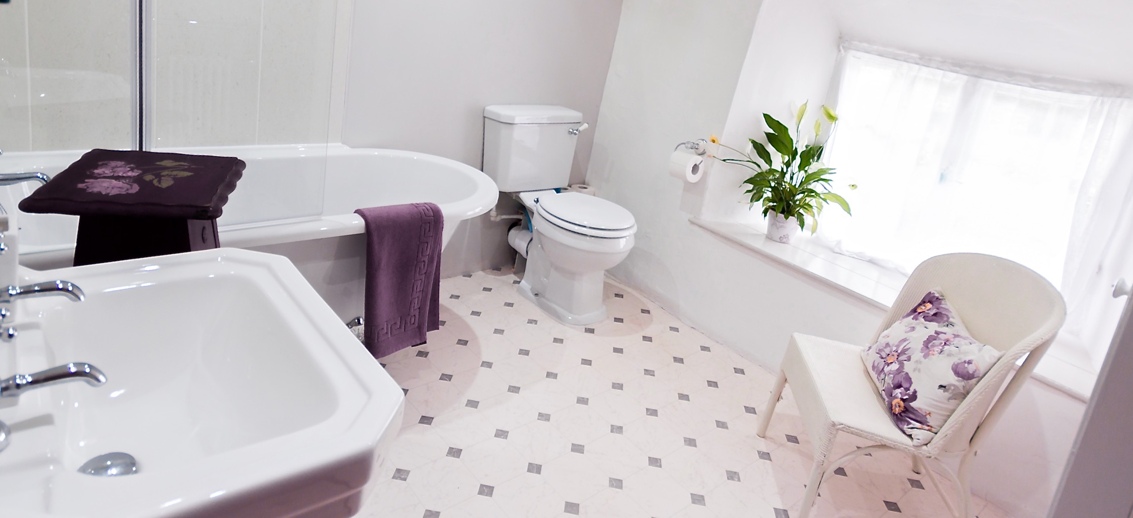 Sun shines through large,net-curtained windows into a a light and airy holiday cottage bathroom with a bath, sink, WC