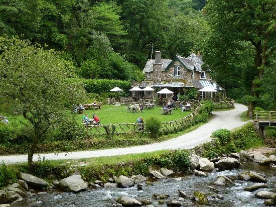 A Moorland river flow around rocks in front of a tearooms and table covered garden surrounded by mature trees in full leaf.