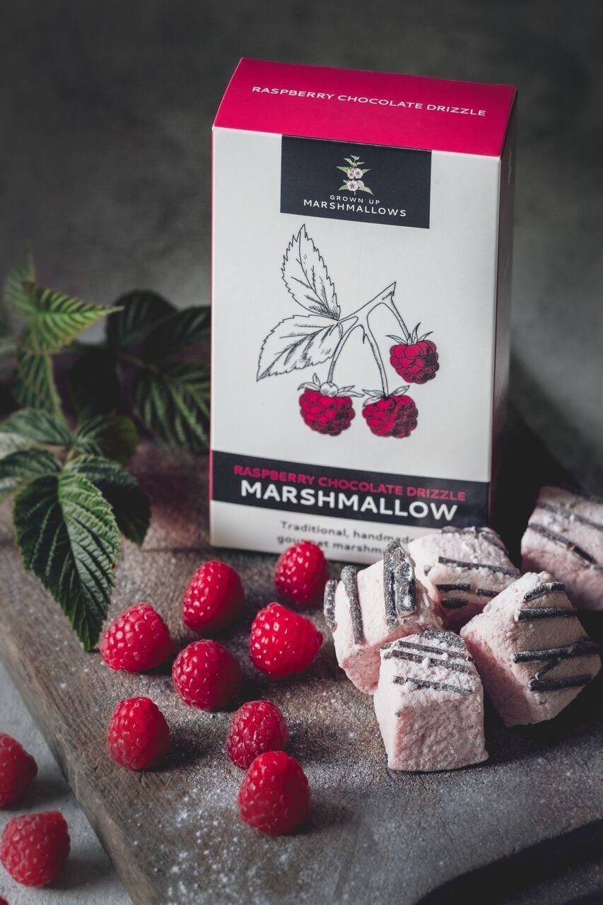 A marshmallow box stands behind squares of marshmallows and some fresh rasperries.