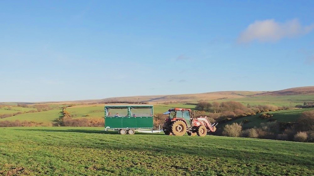 A tractor pulls a trailer in which sit some passengers across an open field with Exmoor hills in the background.