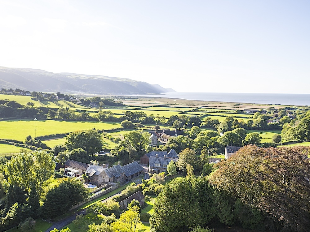 Aerial view of open countryside with a clutch of buildings nestling in the foreground with the Somerset coast on the horizon.