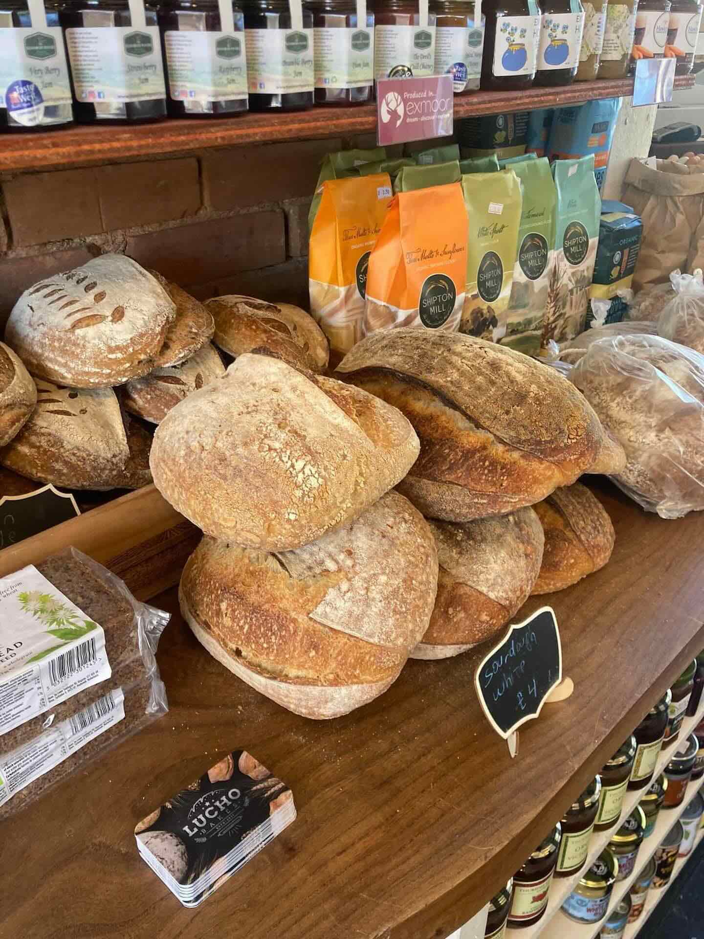 Various types of bread loafs on a shelf in front of other food packets and rows of small jars.