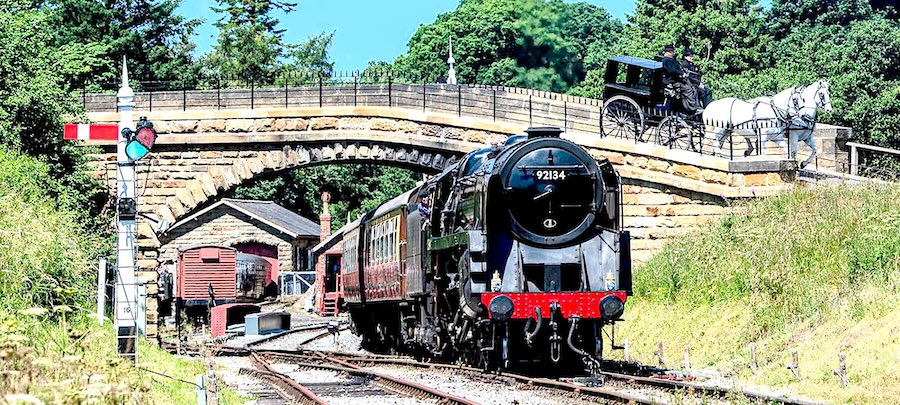 A North York Moors Railway passenger train, hauled by a steam locomotive, pulls away from a station and under a bridge.