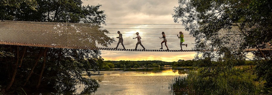 4 children cross a rope bridge over a river are silhouetted against the setting sun.