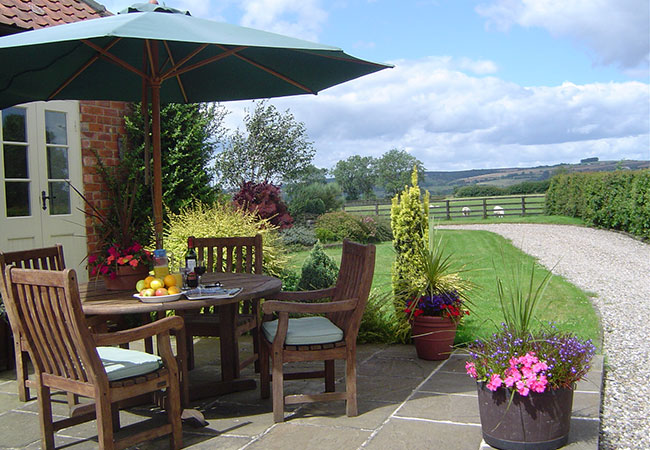 Wooden dining table on a patio outside with a parasol next to a brick cottage. A gravel path leads to fields