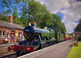 A steam engine waits to depart with its train on a sunny day in Crowcombe 
