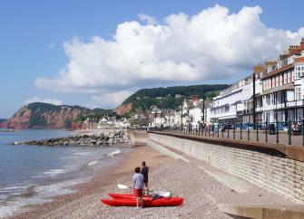 Sidmouth's seafront with the ocean, a sloping pebble beach backed by a long esplanade. Behind this is a row of Georgian hotels and cafes.