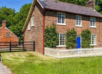 A double-fronted brick-built purpose-built detached cottage surrounded by a white picket fence lawns, and woodland.