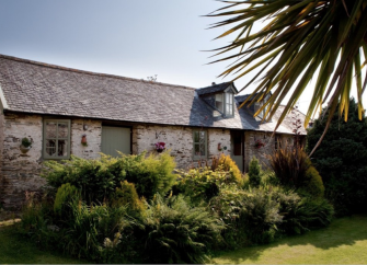 A single-storey stonie0built holiday cottage in North Devon overlooks a spacious lawned garden with shrubs and palm trees.