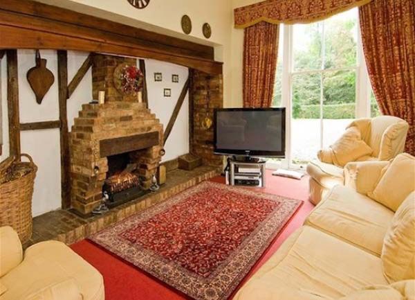 Lounge with a woodburner in a large fireplace facing a 3-piece suite. A TV stands besie tall French windows to a garden.