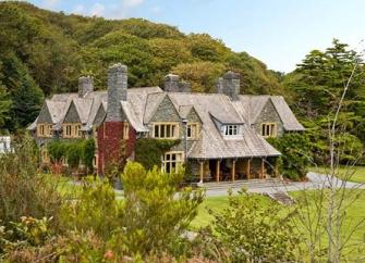 A large slate-roofed house house backed by tall trees overlooks a large lawn.