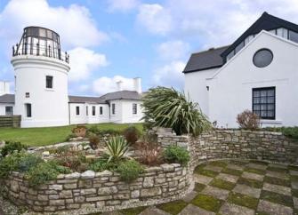 Behind a low stone wall and lawns stands a lighthouse tower and buildings.