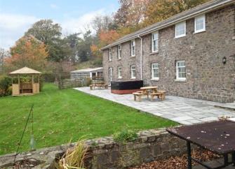 Rear exterior of a long 2-storey cottage overlooking a paved terrace with a hot tub and a lawn.