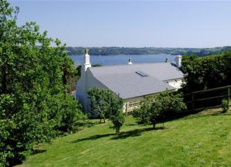 The rooftops of a large holiday cottage surrounded by a sloping lawn and trees with a river estuary in the distance.