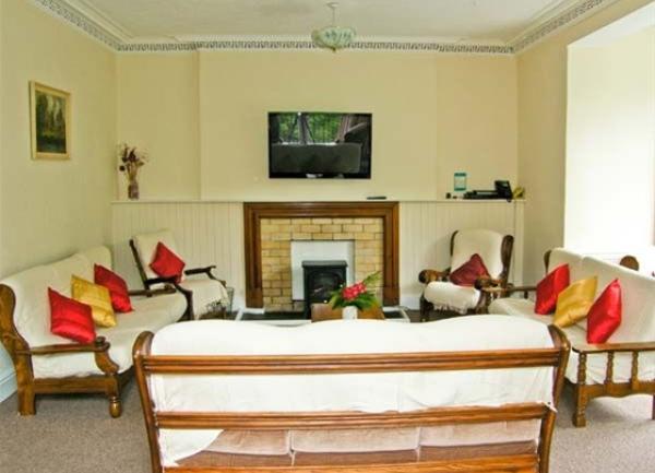 A high-ceilinged, spacious lounge with 3 sofas and two arm chairs facing a woodburner and a wall-mounted TV above.