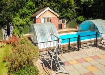 An outdoor swimming pool and terrace with a sun-lounger swing are overlooked by a garden and cottage. 