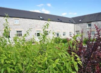 Attached stone barns form an L-shape around a shrub and flower-filled courtyard garden.