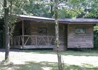 An eco-friendly log cabin with a fenced-in verandah sits in the shade of mature trees.