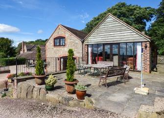 A single storey guilding with a half-timbered apex above a wall of bi-fold glass doors overlooking large patio with outdoor dining furniture.