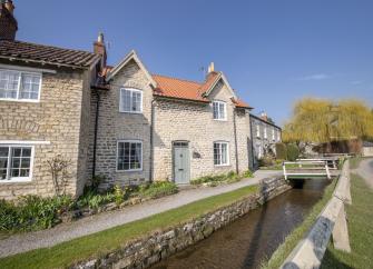 A stone built holiday cottage in Hovingham overlooks a steam running past its frontage on a summer's day.