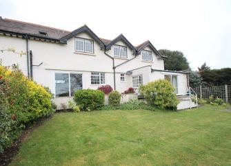 A gable-windowed cottage overlooks a large lawn bordered by bushy shrubs.