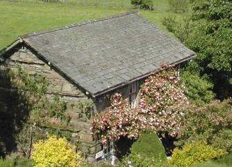A stone built, slate roofed Lake District holiday cottage surrounded by shrubs and small trees with hilly fields in the distance.