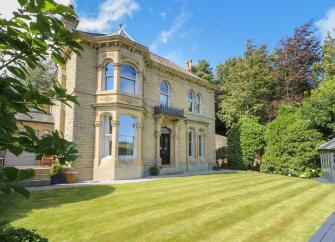 Exterior of a stone-built Victorian Villa overlooking a large lawn bordered by mature shrubs and trees.