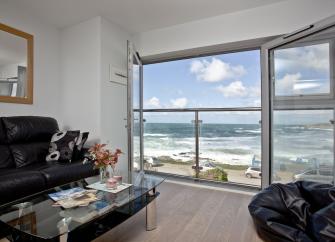 A holiday apartment lounge with floor to ceiling windows beyond which is a view of rhe surf on Ffistral Beach