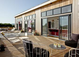 Exterior of a contemporary eco-lodge with large floor-to-ceiling plate glass windows and doors opening onto a wooden deck with a hot tub.