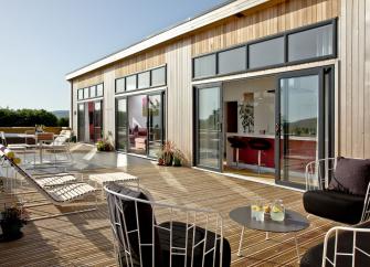 Rxterior of a contemporary eco-loodge with large floor-to-ceiling plate glass windows and doors opening onto a wooden deck with a hot tub.