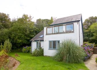 Exterior of a contemporary house in Cornwall with a balcony overlooking a large lawned garden bordered with shrubs and trees.