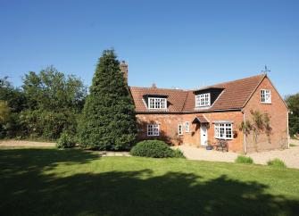 Exterior of an old brick-buily house with dormer windows in the roof. It overlooks a large lawn in which stands a tall, cone-shaped fir tree.
