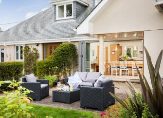 A terrace with chairs and table over look a lawn and mature darden.  Behind the terrace is a large holiday cottage with open floor-to-ceiling French windows.
