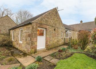 Gable end of a stone built Deerbyshire holiday cottage overlooking a lawn surrounded by mature shrubs.