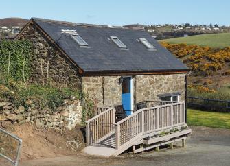 A barn conversion to create a holiday cottage in Abersoch: a wramp leasds to a wooden deck with outdoor seating and an entrance to this stone-built property with rural and sea views.