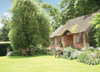 Exterior of s brick-built period garden cottage backed by mature woodland and in front of a lawn with flower-filled borders.