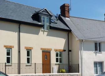 A two-storey converted bakery cottage behind wrought-iron railings in an elevated position above a village street.