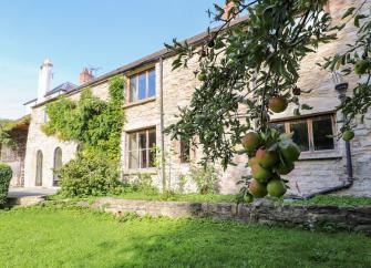 Exterior of a long, stone built Derbyshire holiday cottage overlooking a garden with mature tree.