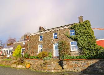 A double fronted, stone-built Welsh farmhouse sits on a flower-filled raised bank above a lane.